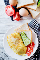 Hearty and tasty breakfast, traditional in the hotel, omelette from chicken eggs with cheese, fresh vegetables - cucumber and tomato, white toast and black coffee on a light wooden background