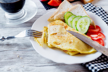 Hearty and tasty breakfast, traditional in the hotel, omelette from chicken eggs with cheese, fresh vegetables - cucumber and tomato, white toast and black coffee on a light wooden background
