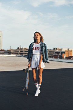 Young female in teenager millenial clothes stands on top rooftop on a sunny warm day in soft sunset light in beautiful shadow, wears hipster socks and longboard