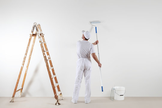 Rear view of painter man looking at blank wall, with paint stick roller, bucket and wooden ladder, isolated on white room