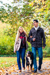 Naklejka na ściany i meble Woman and man with dog having autumn walk on a path covered with foliage