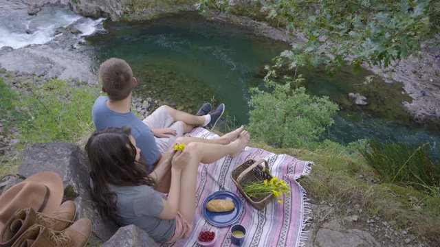 Woman Holds Out Her Hand For Butterfly To Land, She Enjoys A Picnic With Her Boyfriend Overlooking A Beautiful River Below