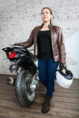 Obraz na płótnie Canvas Full-length portrait of sexy woman in leather jacket standing near motorcycle with helmet in hand, brick wall on background