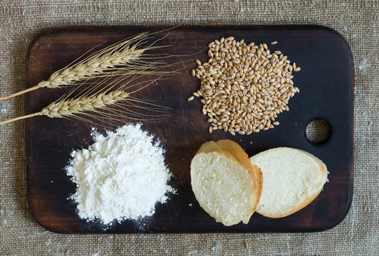 Wheat ears, grains, flour and sliced bread on a kitchen board on a sacking background