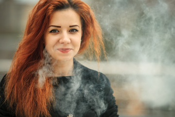 Vape teenager. Young pretty white girl with red curly hair and modern haircut in black dress vaping an electronic cigarette opposite city background.