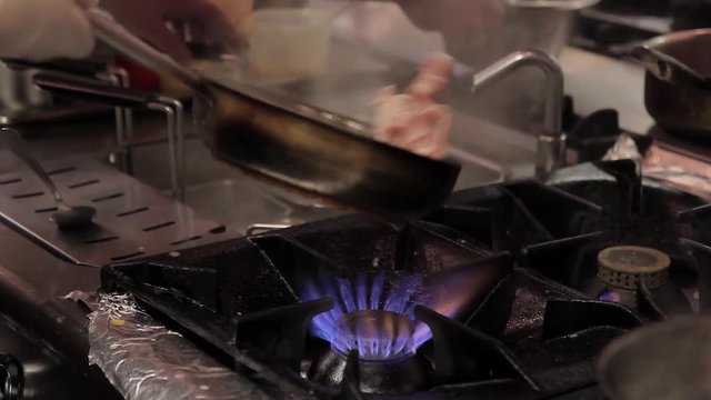 The chef cooks bacon in frying pan on a gas flame in the kitchen of restaurant. The cook in disposable gloves mixes pieces of meat in stainless steel skillet. Dish prepares on the black gas stove. Man