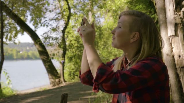 Young Woman Takes Landscape Photos With Her Smartphone In Forest With A Water View