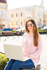 Modern blogger. Side view of beautiful young woman in sunglasses using her laptop and looking away with smile while sitting outdoors sunny street