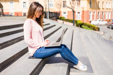 Portrait of a happy young woman sitting on the city stairs and using laptop computer