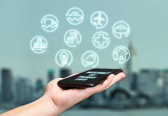 Hand holding mobile with Internet of things (iot) word on screen with icon feature glowing in air with blur city building background,Digital Lifestyle concept