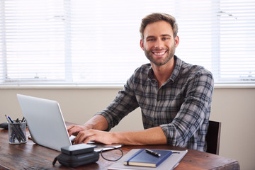 Young caucasian male student smiling at the camera with his hands on the keyboard of his laptop in front of him at his desk.