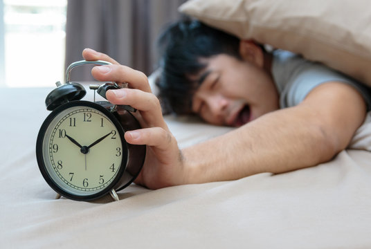 Sleepy young man in bed with eyes closed extending hand to alarm clock