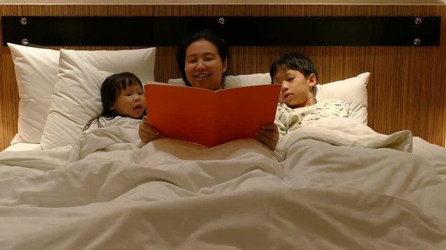 Asian mother reading book for her children on bed