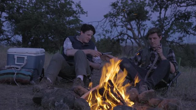 Man Whittles A Piece Of Wood By Campfire, His Friend Asks To Check Out His Knife, He Lets Him Look At It