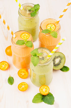 Orange kumquat and green kiwi fruit smoothie in glass jars with straw, mint leaf, cut ripe berry, close up. White wooden board background, vertical.