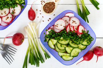 A light vitamin spring - summer salad with fresh cucumbers, radish and green onions, with oil and flax seeds in bowls on a white wooden background. Top view. Dietary, vegan dish. Healthy diet.