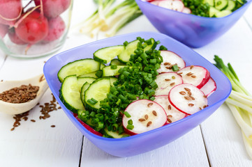 A light vitamin spring - summer salad with fresh cucumbers, radish and green onions, with oil and flax seeds in bowls on a white wooden background. Close up. Dietary, vegan dish. Healthy diet.