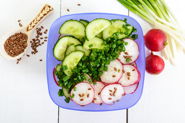 A light vitamin spring - summer salad with fresh cucumbers, radish and green onions, with oil and flax seeds in bowls on a white wooden background. Top view. Dietary, vegan dish. Healthy diet.