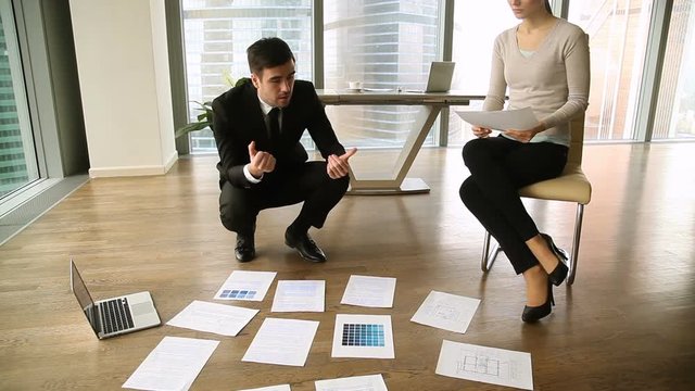 Teamwork of two colleagues in design agency, male designer architect offering variants of flat interior, discussing home remodeling, renovation ideas with female client, papers scattered on the floor