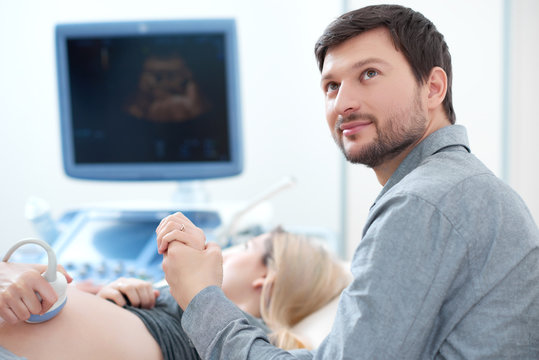 Handsome young  husband carrying about his pregnant wife who diagnosing at ultrasound, screening stomach at hospital. Blonde woman lying in clinic, looking at screen, when doctor showing babies photo.