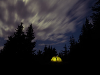 Night landscape with a glowing tourist tent and a night sky surrounded by spruce.