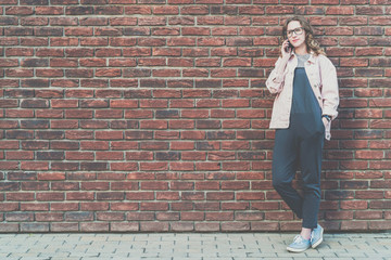 Young attractive hipster woman stands outside on a red brick wall background and talks on her cell phone.