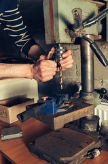 The worker in his garage works on a drilling machine. In the frame, a person's hands