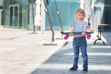 Adorable kid outdoors. Cute pretty child smiling at camera. Casual boy on summer time skating on a skateboard. Holding skateboard like a present