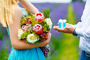 The man's hands stretch out a small gift box with a bow, offering her to marry. She stands in a...