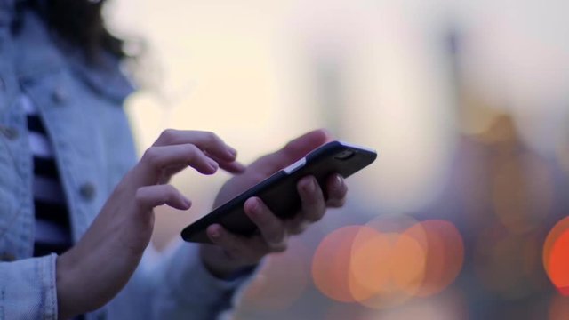 Closeup Of Girl Using Her Smartphone To Text And Send Photos, She Swipes And Zooms In With Her Fingers, City Lights In Background