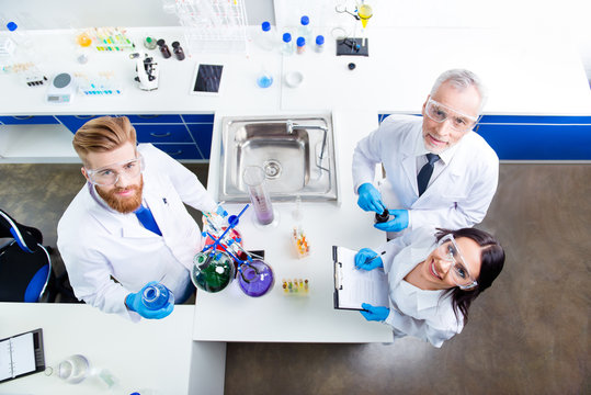 Top up of lab team of three researchers working with reactants and looking up, professor is old and two other workers are young, all are in labcoats