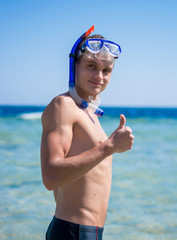 A young man stands near the sea and showing OK sign with his thumb up in the mask for diving