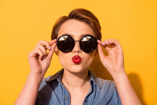 Kiss for you! Fashionable young cute girl in trendy sunglasses sends a kiss against bright yellow background, she holds spectacles with her hands