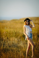 Young pretty woman wearing hat and sunglasses dressed in boho style clothes walking on the nature near river during beautiful sunset