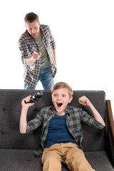 Serious father pointing with finger and excited son sitting on sofa with joystick, family problems concept