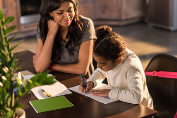 Smiling mother looking at cute little daughter doing homework, homework help concept
