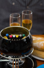 Gourmet Swiss fondue dinner with assorted cheeses and a heated pot of cheese fondue with colorful forks dipping inside of the pot, with white wines cups behind on a wooden table