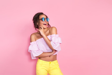 Beautiful girl in colorful clothes wearing sunglasses posing on pink background in studio. - obrazy, fototapety, plakaty