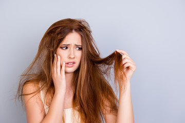 Sad young cute girl is looking at her damaged hair with shock, standing isolated on a pure background, touching her face