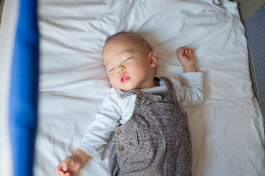 Cute little Asian 1 year old toddler baby boy child sleeping in baby cot, Peaceful kid lying on baby bed while sleeping with windows light at his face, Daytime sleep concept (shallow depth of field)