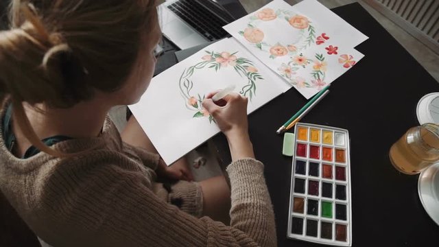 A young woman artist, paints with watercolor paints a water brush flowers, a wreath of green leaves. close up