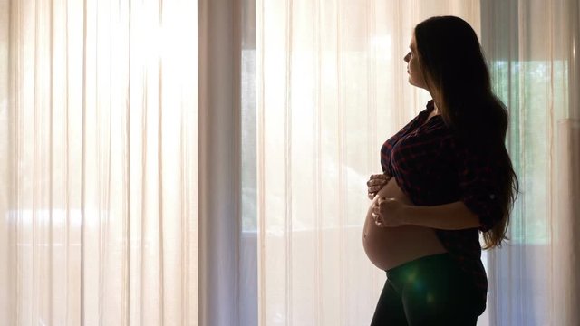 Beautiful pregnant woman standing by the sunny window and touching her tummy in the morning