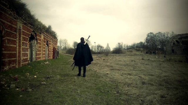 Bearded man dressed in assassin's costume is going along the ruined fortress with a sword  in hand.