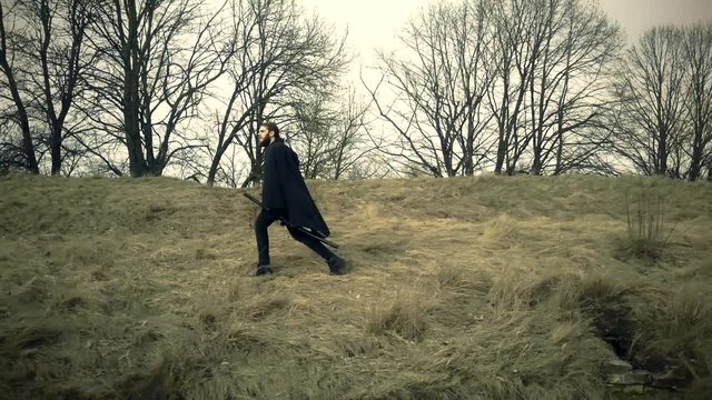 Bearded man dressed in assassin's costume training with a sword on the field.