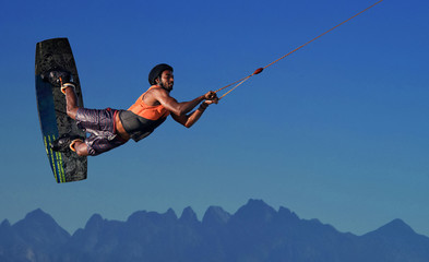 Wake boarding sportsman rider flying high with raley freestyle trick in a deep blue sky and mountains background, extreme water sports and active lifestyle, recreation hobby and fun at leisure time