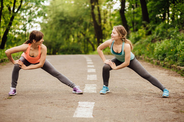 Two fitness women doing stretching exercise before running in green park in summer time.