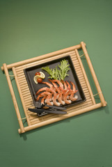 Shrimps with lemon and tomato with fork and knife on black plate on bamboo tray on green background