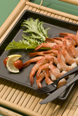 Shrimps with lemon and tomato with fork and knife on black plate on bamboo tray on green background