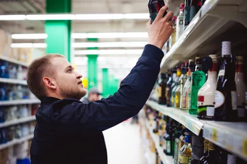 Fototapeten bearded man, In a black jacket, stopped to make my choice in store window, holds a bottle of strong alcohol in his hand, taking from shelf © evgeniykleymenov