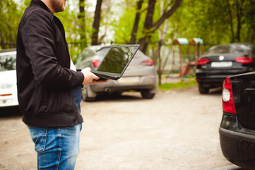 Fototapeta premium man with a laptop in parking lot in yard near car is doing manipulations with cyber system, concept